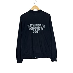 May include: Black long-sleeve jacket with the text "BATHINGAPE CONQUISTA :2001" in white lettering on the back. The jacket is hanging on a wooden hanger against a white background.