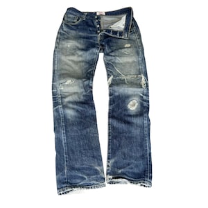 Könnte beinhalten: Eine blaue Jeans aus Denim mit abgenutzten Details, darunter Risse und Ausbleichen. Die Jeans hat einen Knopfverschluss und liegt flach auf einem weißen Hintergrund.