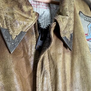 May include: Close-up of a vintage brown leather jacket with a zipper and metal accents. The jacket has a worn, distressed appearance, with visible texture and detailing. The collar features decorative metal emblems.