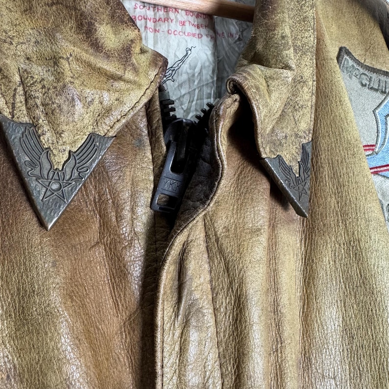 May include: Close-up of a vintage brown leather jacket with a zipper and metal accents. The jacket has a worn, distressed appearance, with visible texture and detailing. The collar features decorative metal emblems.
