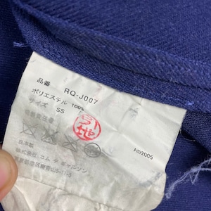 Puede incluir: Una etiqueta blanca con texto negro y un sello rojo. La etiqueta dice "RQ-J007", "Poli&eacute;ster 100%", "Talla SS", "AD2005" y "Hecho en Jap&oacute;n".