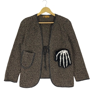 Könnte beinhalten: Eine braune Tweedjacke mit einem schwarzen Skelett-Hand-Patch auf der rechten Tasche. Die Jacke hat einen Bindeverschluss vorne und einen schwarzen Besatz an den Rändern.