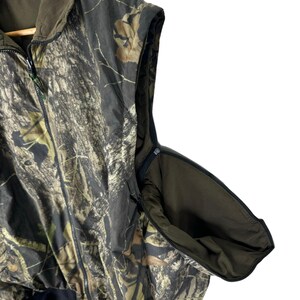 May include: A sleeveless camouflage hunting vest with a zip closure and a hood. The vest has a large pocket on the side.