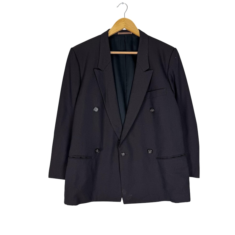 Op de afbeelding: Een zwarte double-breasted blazer hangt aan een houten hanger. De blazer heeft twee rijen knopen, een gekerfde revers en twee voorzakken. De jas is gemaakt van een donkere stof.