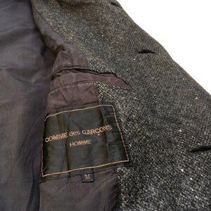 May include: A black label with white text that reads "COMME des GARCONS HOMME" with a size "M" label below. The label is attached to a gray tweed jacket.
