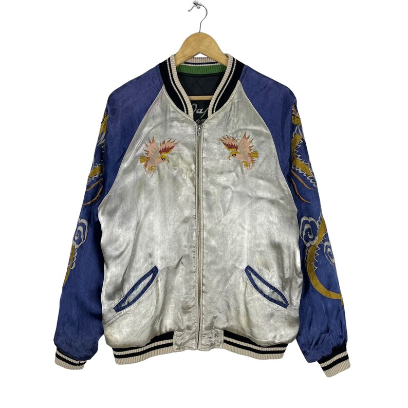 May include: A silver satin bomber jacket with blue sleeves. The sleeves feature embroidered dragons and clouds. The jacket has a zipper closure and a ribbed collar, cuffs, and hem.