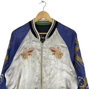 May include: A vintage satin bomber jacket with a blue and white color scheme. The jacket features embroidered eagles on the front and a dragon design on the sleeves. The jacket has a zipper closure and a ribbed collar.