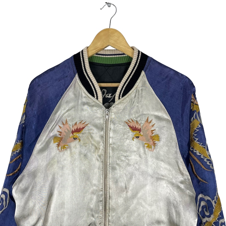 May include: A vintage satin bomber jacket with a blue and white color scheme. The jacket features embroidered eagles on the front and a dragon design on the sleeves. The jacket has a zipper closure and a ribbed collar.