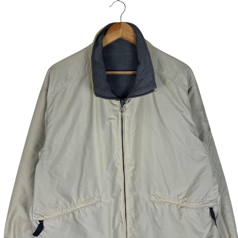 Puede incluir: Chaqueta reversible beige con cremallera completa y cuello gris oscuro. La chaqueta tiene dos bolsillos delanteros y est&aacute; colgada de una percha de madera. La chaqueta est&aacute; hecha de un material ligero.