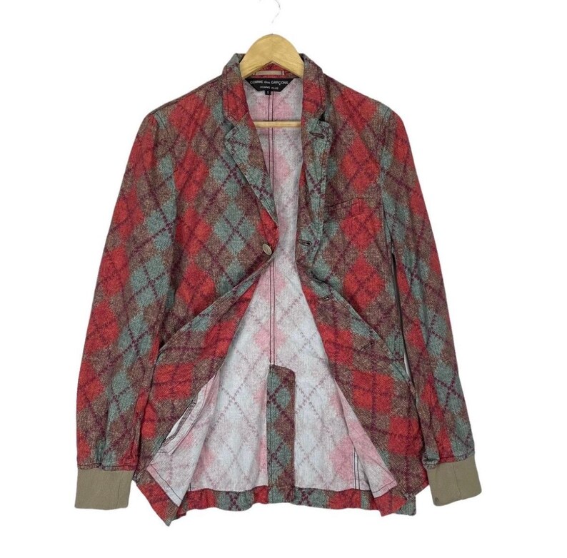 May include: A red, green, and brown plaid blazer with a single-button closure. The blazer has long sleeves with brown cuffs and is hanging on a wooden hanger. The interior lining is a lighter plaid pattern.