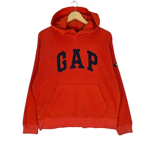 May include: A red hooded sweatshirt with a navy blue "GAP" logo on the front. The sweatshirt has a kangaroo pocket and a drawstring hood.