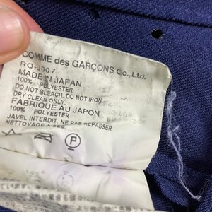 Puede incluir: Una etiqueta blanca con texto negro sobre un fondo de tela azul marino. La etiqueta dice "COMME des GAR&Ccedil;ONS Co., Ltd. RQ-J007 MADE IN JAPAN 100% POLYESTER DO NOT BLEACH. DO NOT IRON DRY CLEAN ONLY. FABRIQUE AU JAPON 100% POLYESTER JAVEL INTERDIT. NE PAS REPASSER. NETTOYAGE A SEC."