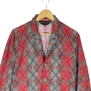 May include: A red, green, and brown argyle patterned blazer with a two-button closure. The blazer has a notched lapel and a breast pocket. The label reads "COMME des GAR&Ccedil;ONS HOMME PLUS".
