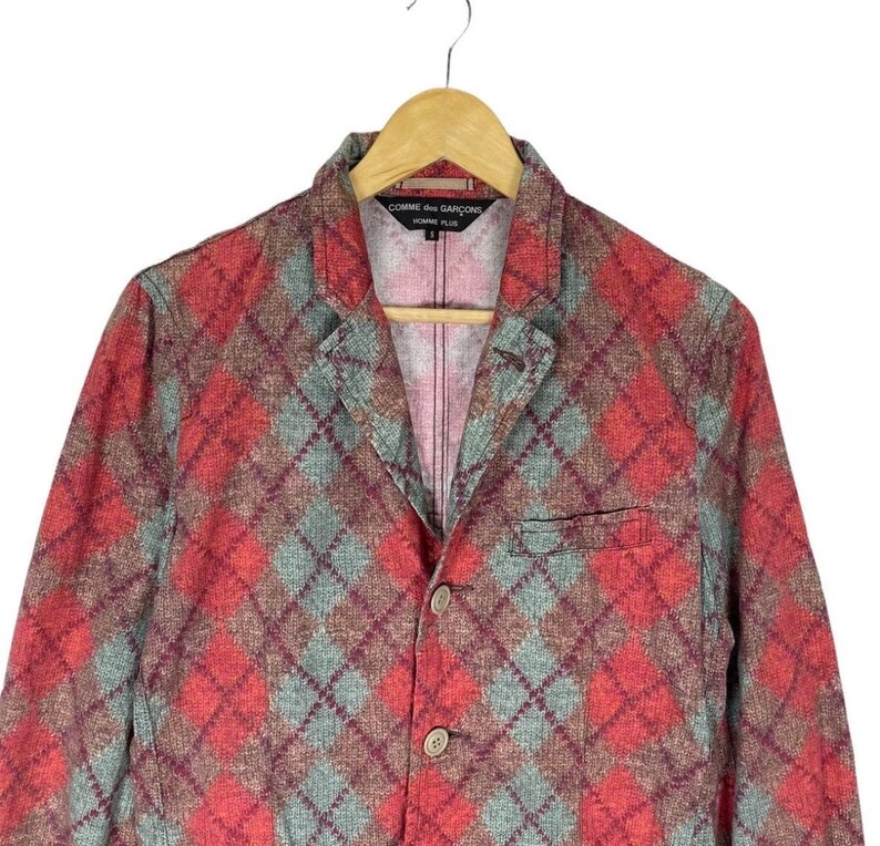 May include: A red, green, and brown argyle patterned blazer with a two-button closure. The blazer has a notched lapel and a breast pocket. The label reads "COMME des GAR&Ccedil;ONS HOMME PLUS".