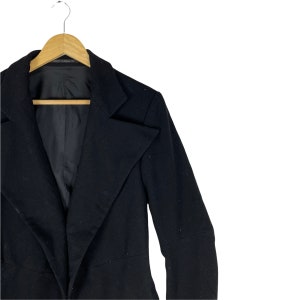 May include: A black blazer with a long, pointed lapel and long sleeves. The blazer is hanging on a wooden hanger.