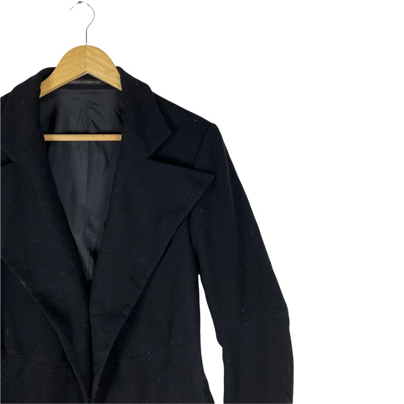 May include: A black blazer with a long, pointed lapel and long sleeves. The blazer is hanging on a wooden hanger.