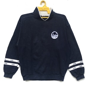 May include: A navy blue sweatshirt with white and blue striped cuffs. The sweatshirt has a mock neck and a small embroidered logo on the front that says "ellesse dal 1959".