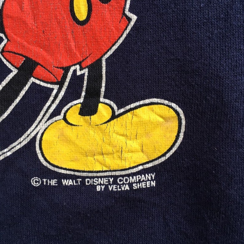 K&ouml;nnte beinhalten: Ein marineblaues Sweatshirt mit einem Cartoon-Bild von Mickey Mouse. Das Sweatshirt hat den Text "&copy; THE WALT DISNEY COMPANY BY VELVA SHEEN" unten aufgedruckt.