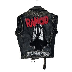 May include: Black leather vest with the word "RANCID" in red lettering across the back. A black and white graphic is below the word "RANCID". The text "AND OUT COME THE WOLVES" is at the bottom of the vest.