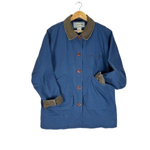May include: A blue jacket with a brown corduroy collar and cuffs. The jacket has four large pockets, brown buttons, and a small pocket on the chest. The jacket is hanging on a wooden hanger.