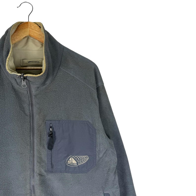 Puede incluir: Chaqueta de forro polar gris con cremallera completa y cuello alto. La chaqueta tiene un bolsillo azul oscuro en el pecho con las letras "ACG" y un gr&aacute;fico. El interior del cuello es de color beige. La chaqueta est&aacute; colgada en una percha de madera.