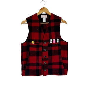 Puede incluir: Un chaleco de lana a cuadros rojos y negros con cuello en V y cierre de botones. El chaleco tiene dos bolsillos, uno con un objeto amarillo y el otro con tres encendedores. El chaleco est&aacute; colgado de una percha de madera.