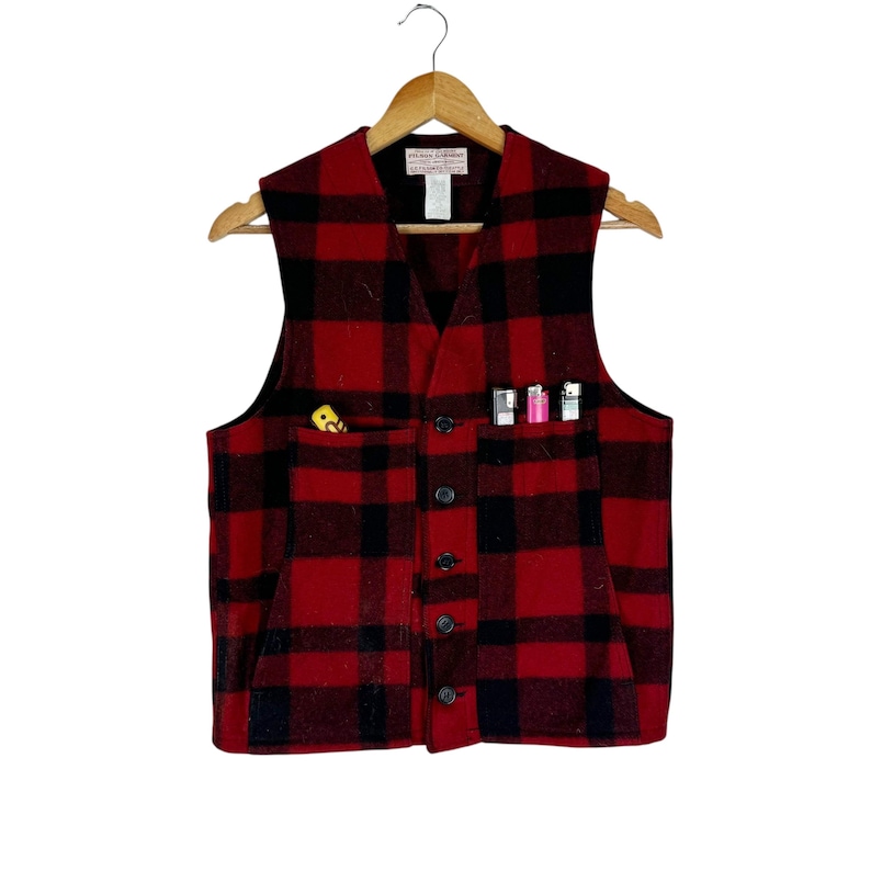 Puede incluir: Un chaleco de lana a cuadros rojos y negros con cuello en V y cierre de botones. El chaleco tiene dos bolsillos, uno con un objeto amarillo y el otro con tres encendedores. El chaleco est&aacute; colgado de una percha de madera.