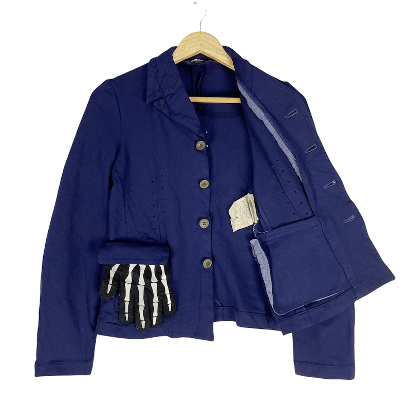 Puede incluir: Una chaqueta azul marino con un dise&ntilde;o de mano de esqueleto en el bolsillo. La chaqueta tiene cierre de botones y un solo bolsillo en el lado izquierdo. La chaqueta est&aacute; abierta para mostrar el forro.