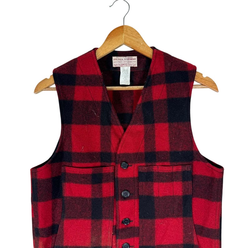 Puede incluir: Un chaleco de lana a cuadros rojos y negros con cuello en V y cierre de botones. El chaleco tiene dos bolsillos delanteros y una peque&ntilde;a etiqueta en el cuello. Cuelga de una percha de madera.