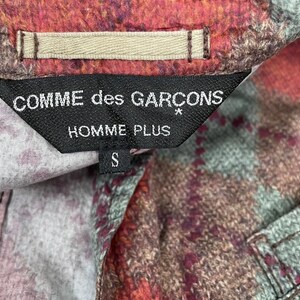 May include: A close-up of a shirt label displaying "COMME des GAR&Ccedil;ONS HOMME PLUS" in white on a black tag. The shirt features a colourful plaid pattern in red, brown, and green. The size "S" is also visible on the label.