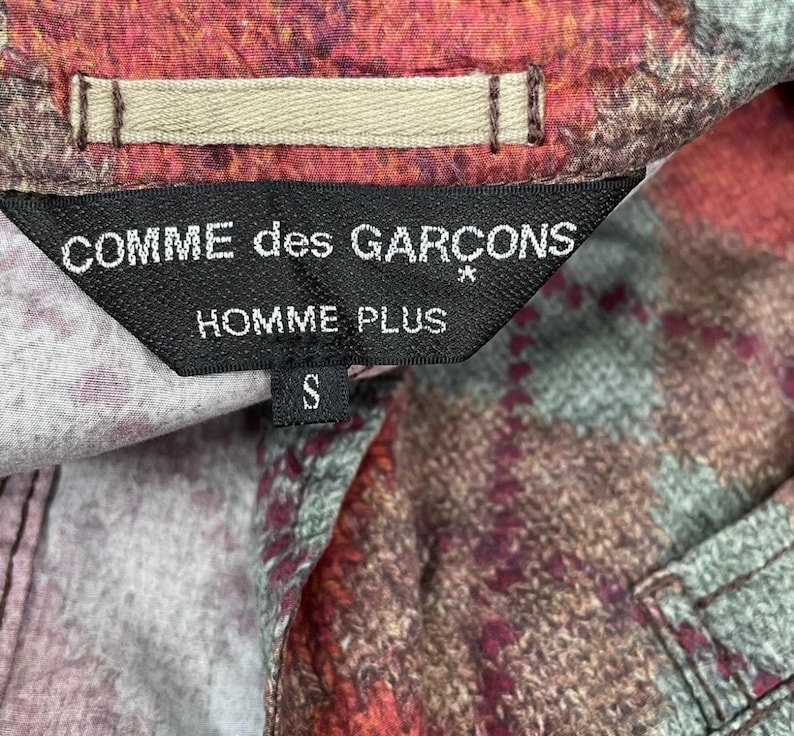 May include: A close-up of a shirt label displaying "COMME des GAR&Ccedil;ONS HOMME PLUS" in white on a black tag. The shirt features a colourful plaid pattern in red, brown, and green. The size "S" is also visible on the label.
