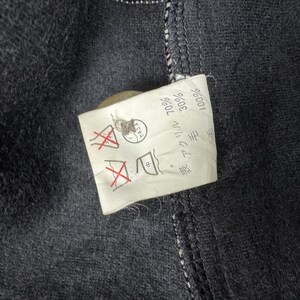 Op de afbeelding: Close-up van een donkergrijze stof met een wit label eraan. Het label toont verzorgingssymbolen en Japanse tekst die de samenstelling van de stof aangeeft: 70% acryl, 30% wol. De stof heeft zichtbare stiksels en een knoop.