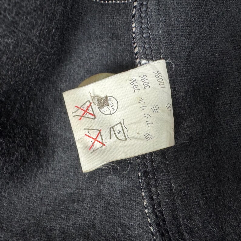 Op de afbeelding: Close-up van een donkergrijze stof met een wit label eraan. Het label toont verzorgingssymbolen en Japanse tekst die de samenstelling van de stof aangeeft: 70% acryl, 30% wol. De stof heeft zichtbare stiksels en een knoop.