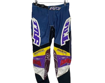 Op de afbeelding: Blauwe, zwarte, witte, paarse en gele motocrossbroek met het woord "TROY" bedrukt op de zijkanten. De broek heeft een zwarte riem en een witte rits.