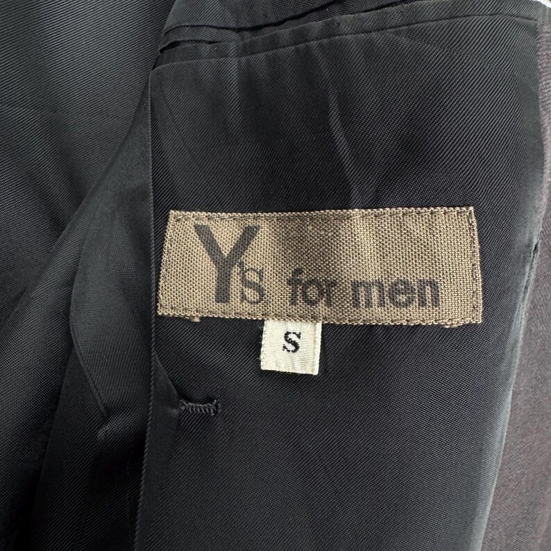 Op de afbeelding: Detail van de binnenkant van een donkergrijze blazer. Een rechthoekig label met de tekst "Y's for men" is zichtbaar, samen met een kleiner label met de letter "S". De voering van de blazer is donkerder grijs.