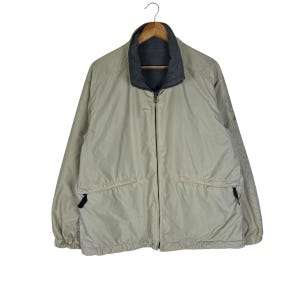 Puede incluir: Chaqueta reversible de color beige neutro, con cuello e interior gris oscuro. La chaqueta tiene cremallera completa, dos bolsillos delanteros y un dise&ntilde;o sutil en la manga. Cuelga de una percha de madera.