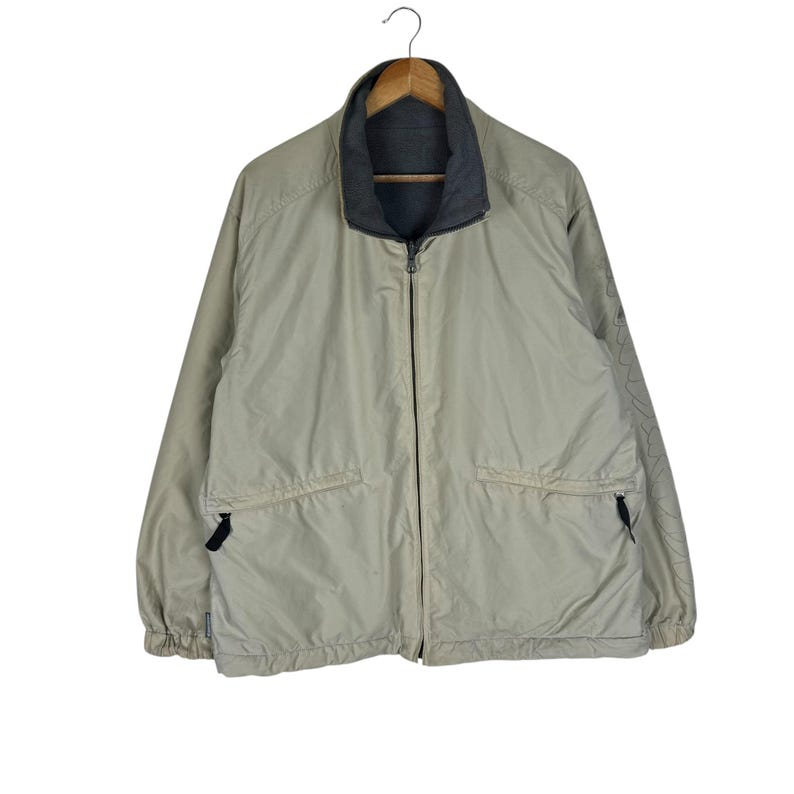 Puede incluir: Chaqueta reversible de color beige neutro, con cuello e interior gris oscuro. La chaqueta tiene cremallera completa, dos bolsillos delanteros y un dise&ntilde;o sutil en la manga. Cuelga de una percha de madera.