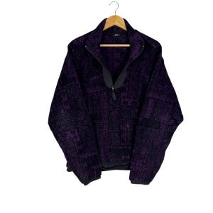 Puede incluir: Un jersey de forro polar morado y negro con un estampado geométrico. La chaqueta tiene una cremallera negra y un cuello alto. La prenda está colgada de una percha de madera sobre un fondo blanco.