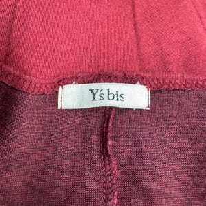 Op de afbeelding: Close-up van een bordeauxrood kledingstuk, mogelijk een top of rok, met een zichtbare naad en een label. Het label leest "Y's bis" in een eenvoudige lettertype, wat duidt op een merk. De stof lijkt een zachte tricot te zijn.
