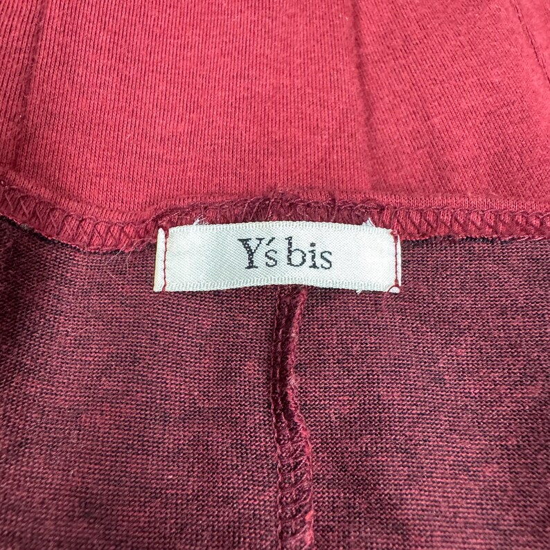 Op de afbeelding: Close-up van een bordeauxrood kledingstuk, mogelijk een top of rok, met een zichtbare naad en een label. Het label leest "Y's bis" in een eenvoudige lettertype, wat duidt op een merk. De stof lijkt een zachte tricot te zijn.