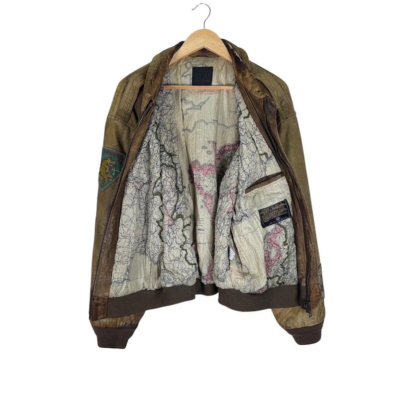 May include: A brown leather bomber jacket with a map-printed lining. The jacket features a zip-up front, ribbed cuffs, and a patch on the upper left sleeve. The interior lining displays a detailed map design with red and green accents.