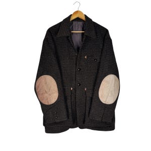 Puede incluir: Chaqueta de tweed marrón oscuro con coderas color beige, colgada de una percha de madera. La chaqueta tiene cuello, botones y dos bolsillos delanteros. Chaqueta de estilo clásico.