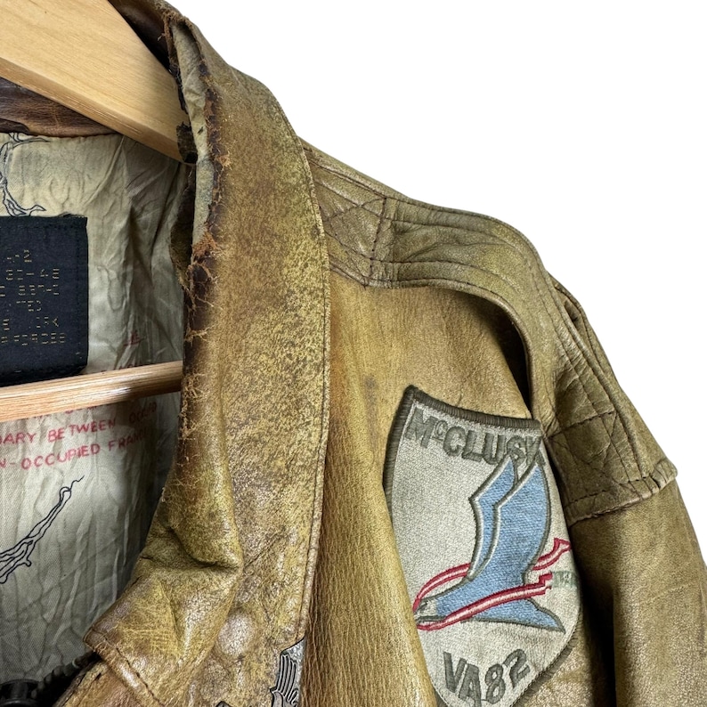 May include: Close-up of a vintage, distressed brown leather jacket. Features a patch with the name "McClusky" and a blue bird design with red accents. The jacket shows signs of wear, with visible texture and a worn appearance.