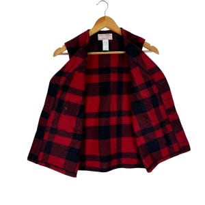 Puede incluir: Un chaleco a cuadros rojos y negros colgado de una percha de madera. El chaleco est&aacute; abierto, revelando el forro interior con el mismo dise&ntilde;o a cuadros. La prenda tiene cuello y parece estar hecha de un material grueso y c&aacute;lido.