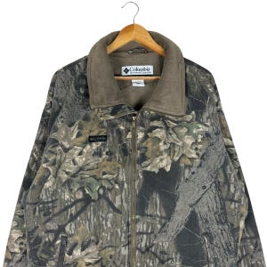 May include: A Columbia Sportswear camouflage jacket with a full zip and a brown fleece collar. The jacket has a woodland camouflage pattern in shades of brown, green, and grey. The Columbia logo is visible on the chest and collar.