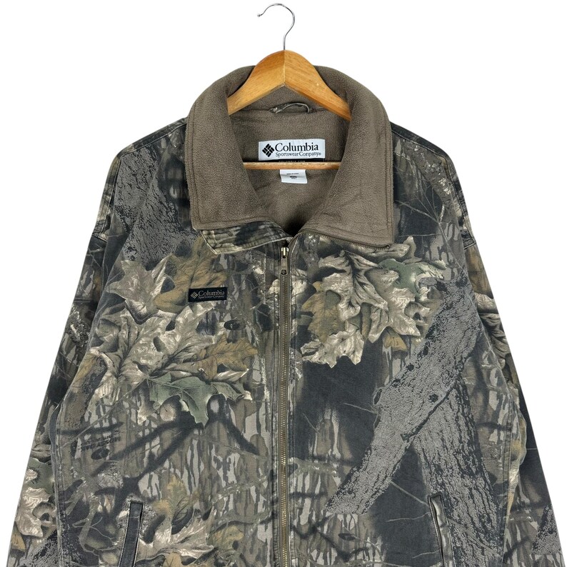 May include: A Columbia Sportswear camouflage jacket with a full zip and a brown fleece collar. The jacket has a woodland camouflage pattern in shades of brown, green, and grey. The Columbia logo is visible on the chest and collar.