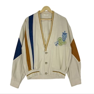 Könnte beinhalten: Ein beigefarbener Cardigan mit V-Ausschnitt und Knopfleiste. Der Cardigan hat ein gestreiftes Muster an den Ärmeln und ein buntes Stickmuster auf der Vorderseite.