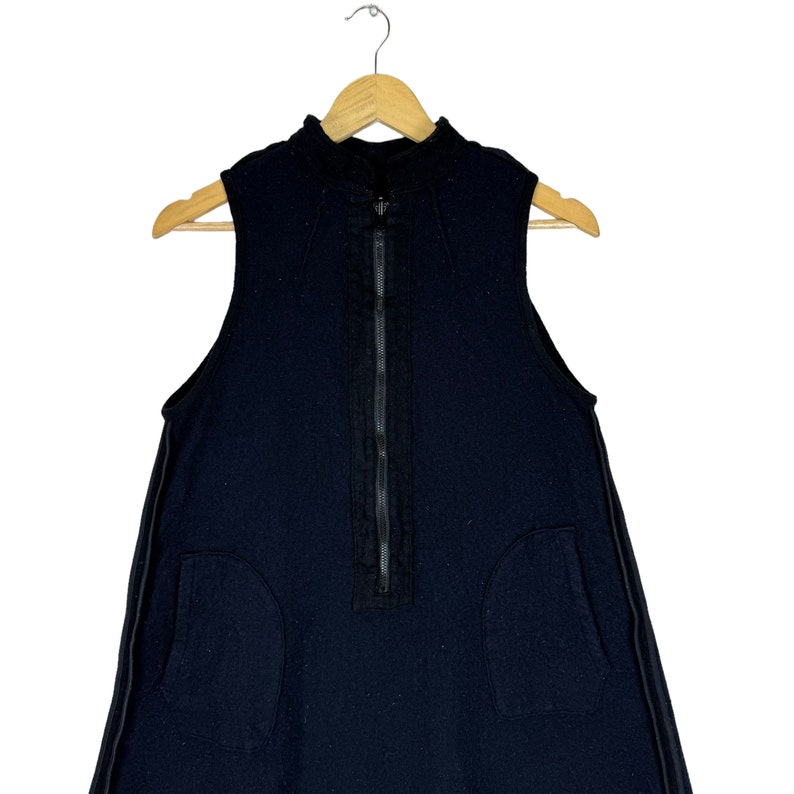 K&ouml;nnte beinhalten: Ein schwarzes, &auml;rmelloses Kleid mit einem Rei&szlig;verschluss an der Vorderseite und zwei Taschen an den Seiten. Das Kleid hat einen hohen Ausschnitt und eine lockere Passform.