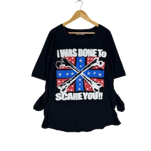 May include: Black t-shirt with the text "I WAS BONE TO SCARE YOU!!" and a graphic of crossed bones over a red and blue design with stars. The shirt is hanging on a wooden hanger.