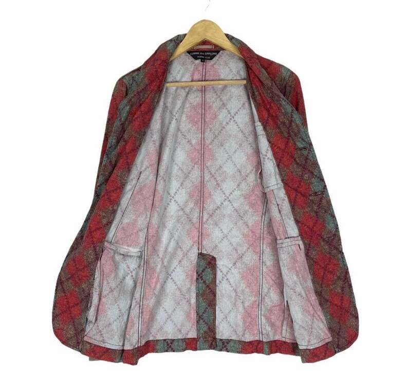 May include: A red, grey, and green plaid jacket with a diamond pattern lining. The jacket is open, revealing the interior lining and pockets. It hangs on a wooden hanger.